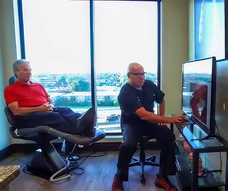Doctor Tom Hall in black scrubs discusses an X-ray on a screen with a seated patient in red. They’re in an office with a large window, creating a professional yet relaxed atmosphere.