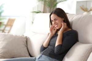 Woman sitting on sofa, grimacing in pain due to TMJ disorder symptoms 